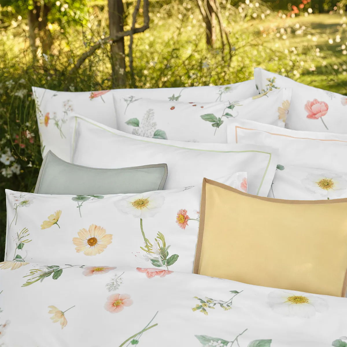 Detailaufnahme von verschiedenen Kissen der Schlossberg Bettwäsche Zinnia, darunter das Solene Kissen in gelb mit braunem Rand, umgeben von weiteren Kissen mit floralen Mustern auf weißem Hintergrund.