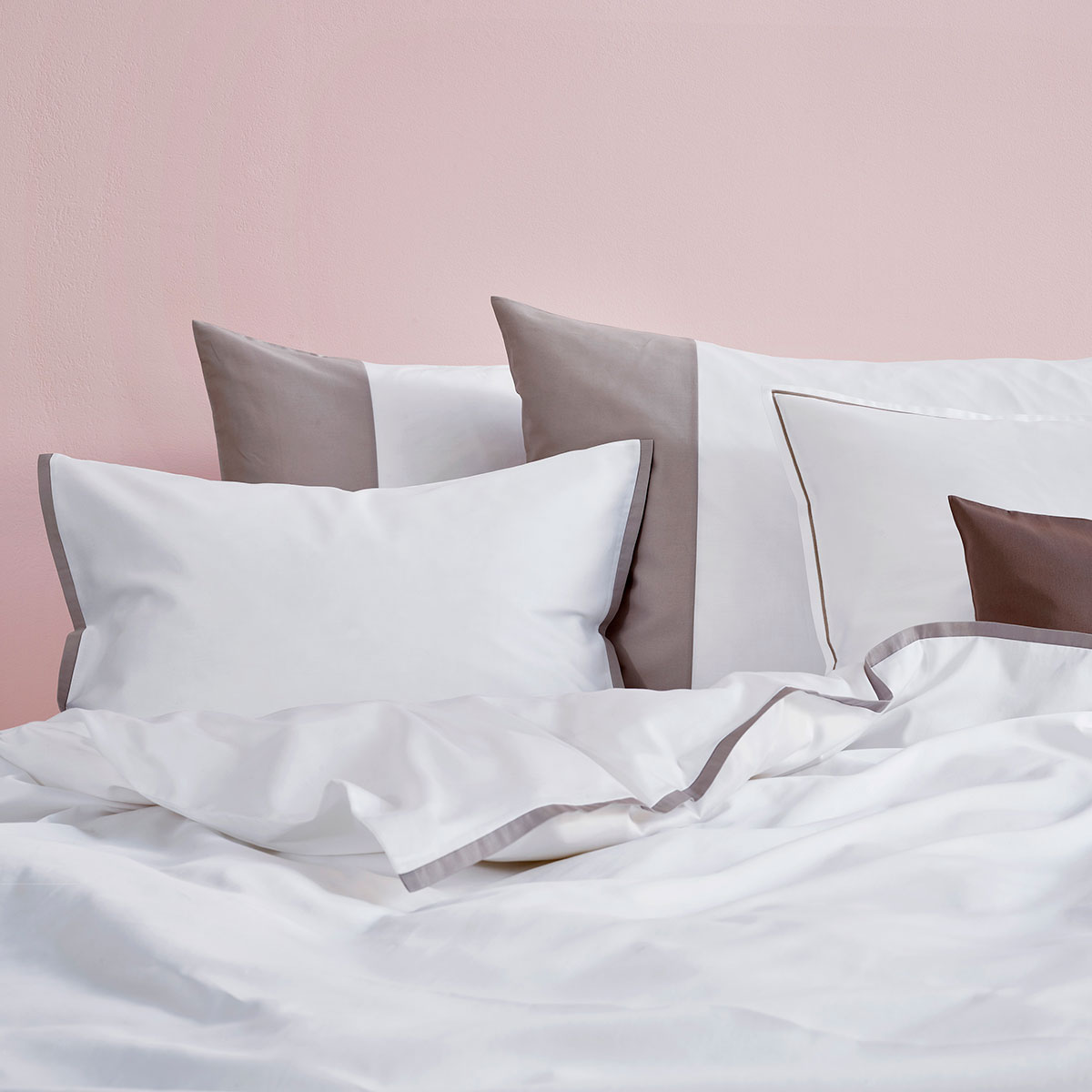 Bettwäsche Allen und Kissen vor rosa Wand Ein Bett mit weisser und grauer Bettwäsche vor einer rosa Wand. Die Kissen sind in verschiedenen Grössen und Farben, hauptsächlich weiss und grau, dekorativ angeordnet.
