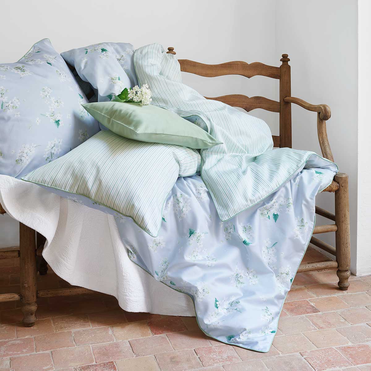 Bettwäsche April im rustikalen Stil Ein rustikales Zimmer mit einer Holzbank, auf der Kissen und Decken der Bettwäsche April in hellblau mit floralem Muster und grünen Streifen liegen.