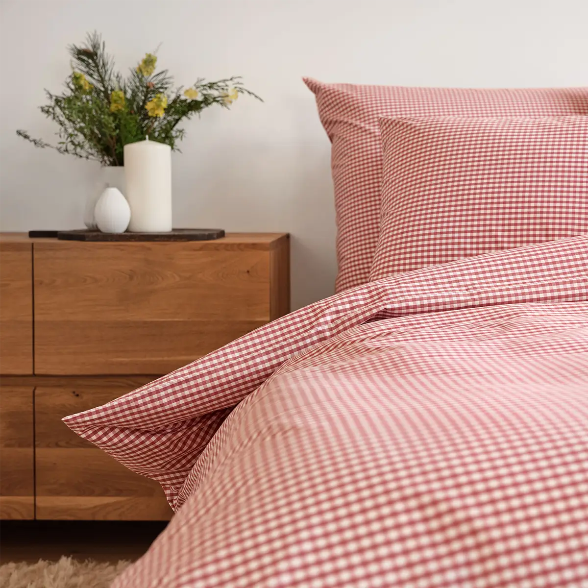 Schlossberg Bettwäsche Andrin Rouge Schlafzimmer Frontal Eine Nahaufnahme der Schlossberg Bettwäsche Andrin Rouge im Schlafzimmer, das mit einem warmen Holzsideboard und einer Vase mit frischen Blumen dekoriert ist. Das rote Karomuster der Bettwäsche verleiht dem Raum eine gemütliche und einladende Atmosphäre
