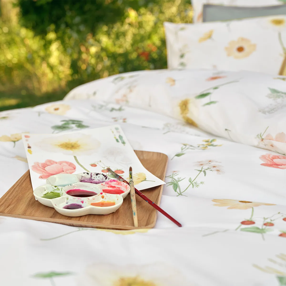 Detailaufnahme der Schlossberg Bettwäsche Zinnia, auf der eine Wasserfarbenpalette und Pinsel auf einem Holzbrett abgebildet sind. Neben der Palette liegt ein gemaltes Blatt mit floralen Motiven, die den Blumen auf der Bettwäsche ähneln.