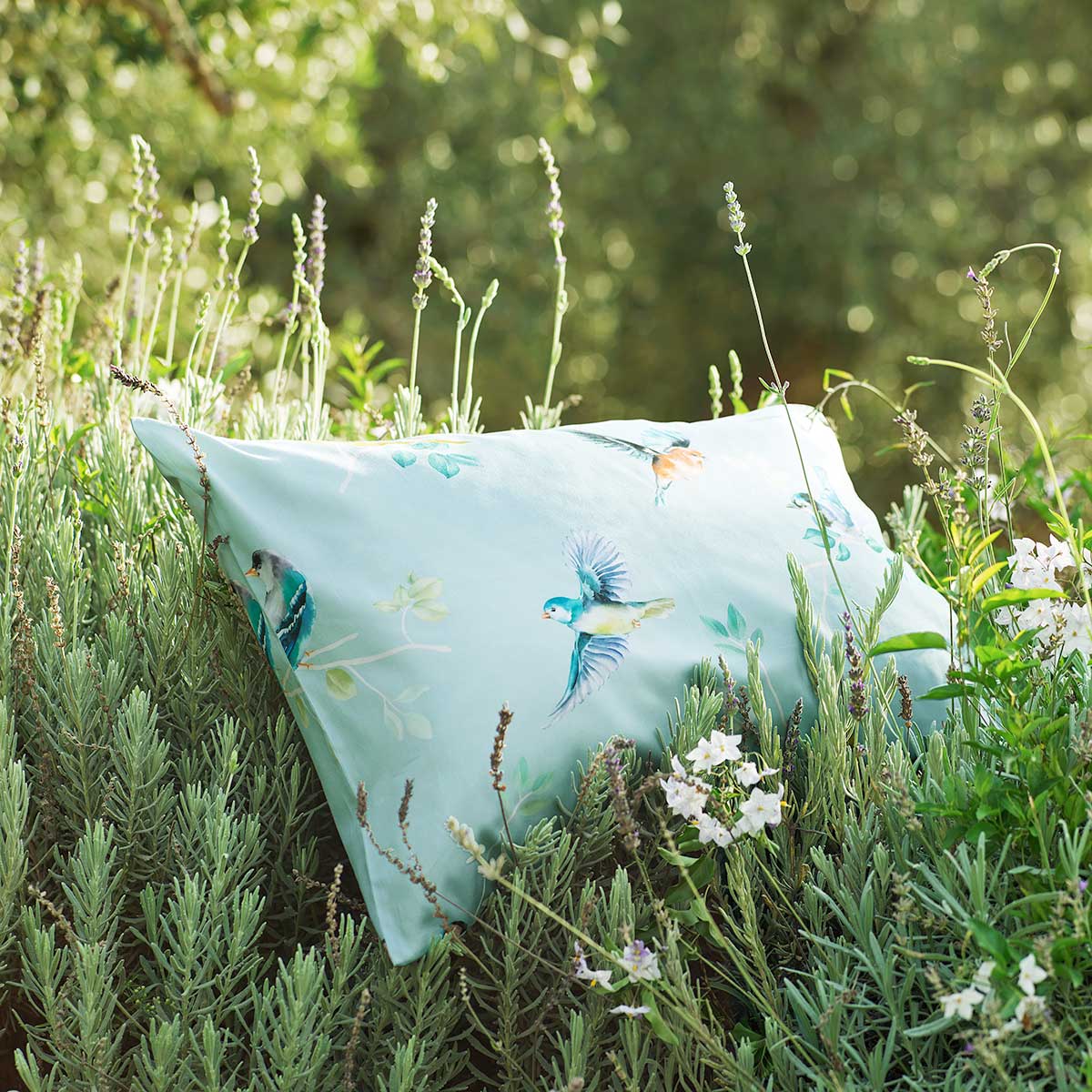 Ein Kissen der Bettwäsche Chirp liegt im Grünen, umgeben von Lavendel und weißen Blumen.