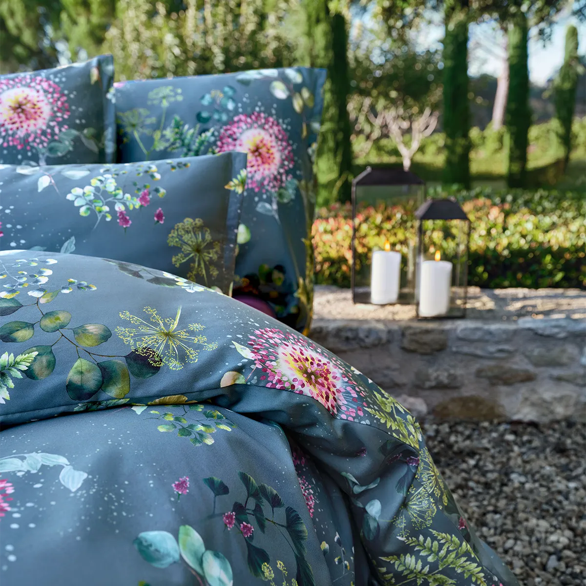 Detailaufnahme der Schlossberg Bettwäsche Nele mit floralen Mustern auf tiefblauem Stoff. Zarte Blüten in Pink, Grün und Gelb schaffen eine elegante Atmosphäre, ergänzt durch sanft leuchtende Laternen im Hintergrund für ein stimmungsvolles Ambiente.