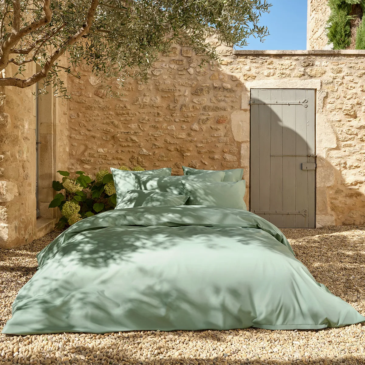Ein stilvoll arrangiertes Bett im Freien mit der Schlossberg Bettwäsche Ben Leaf in einem sanften, beruhigenden Grünton. Die hochwertige Bettwäsche fügt sich harmonisch in die mediterrane Kulisse mit Natursteinmauern und Olivenbaum-Schatten ein.