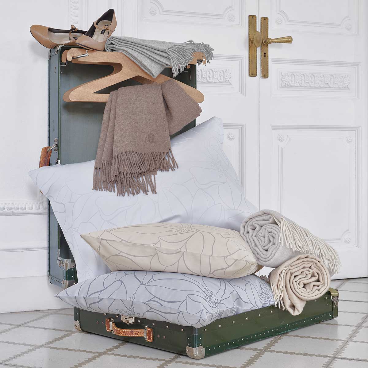 Magnolia Kissen und Decken mit Schal auf einem Koffer Mehrere Magnolia Kissen und Decken in Grau und Beige, dekoriert mit Schals und platziert auf einem offenen Koffer.