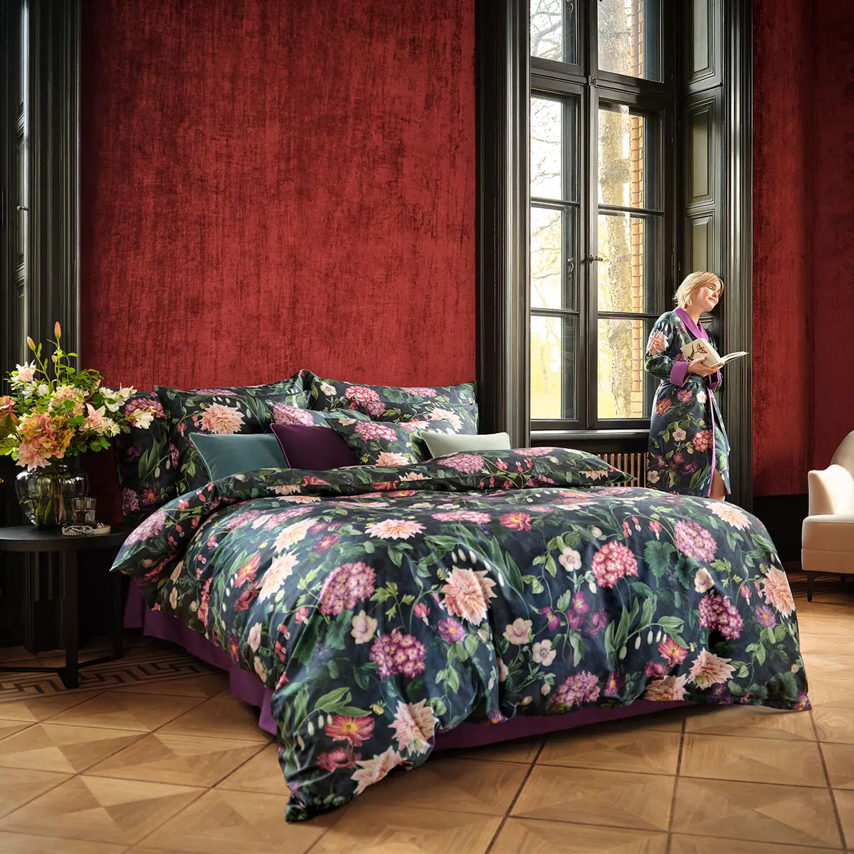 Elegantes Schlafzimmer mit Ophelia Bettwäsche Ein grosszügiges Schlafzimmer mit hohen Fenstern und einer prächtigen, dunkelblauen Ophelia Bettwäsche, die ein florales Muster zeigt. Eine Frau steht am Fenster und liest ein Buch.