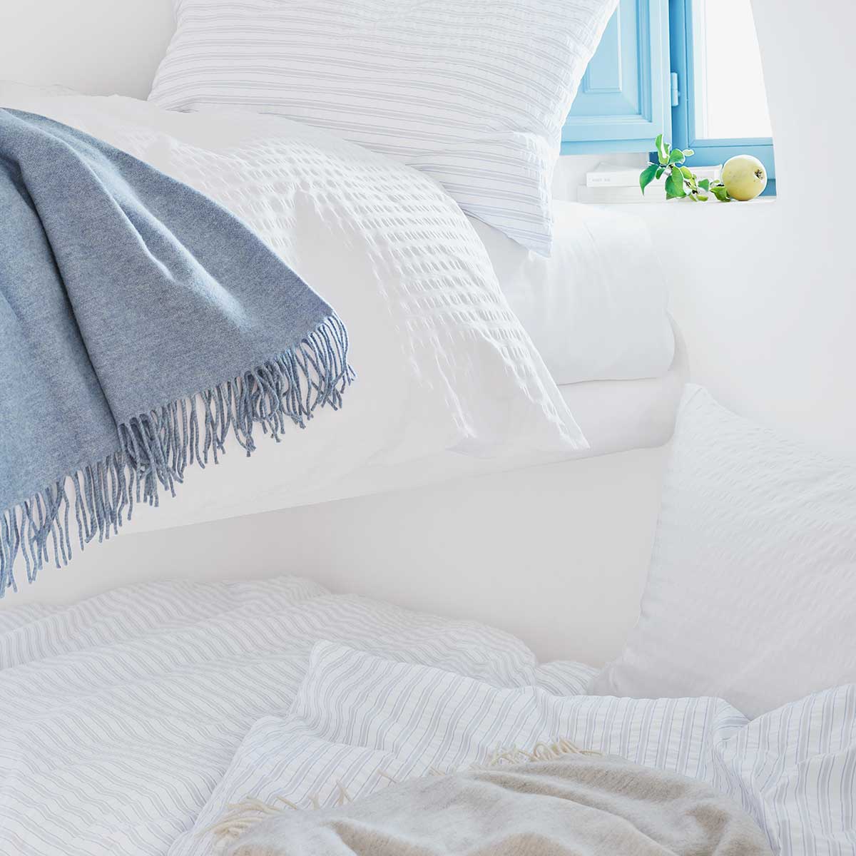 Bettwäsche Dean im Schlafzimmer Bett mit Bettwäsche Dean, in Weiss und blau-weiss gestreift, dekoriert mit einer blauen Decke und umgeben von einem hellen, luftigen Schlafzimmerinterieur.