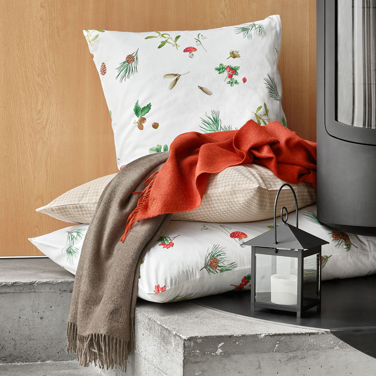 Kissen Hazel mit Waldmuster neben einem roten Schal und einer Laterne, ideal für eine gemütliche Atmosphäre.