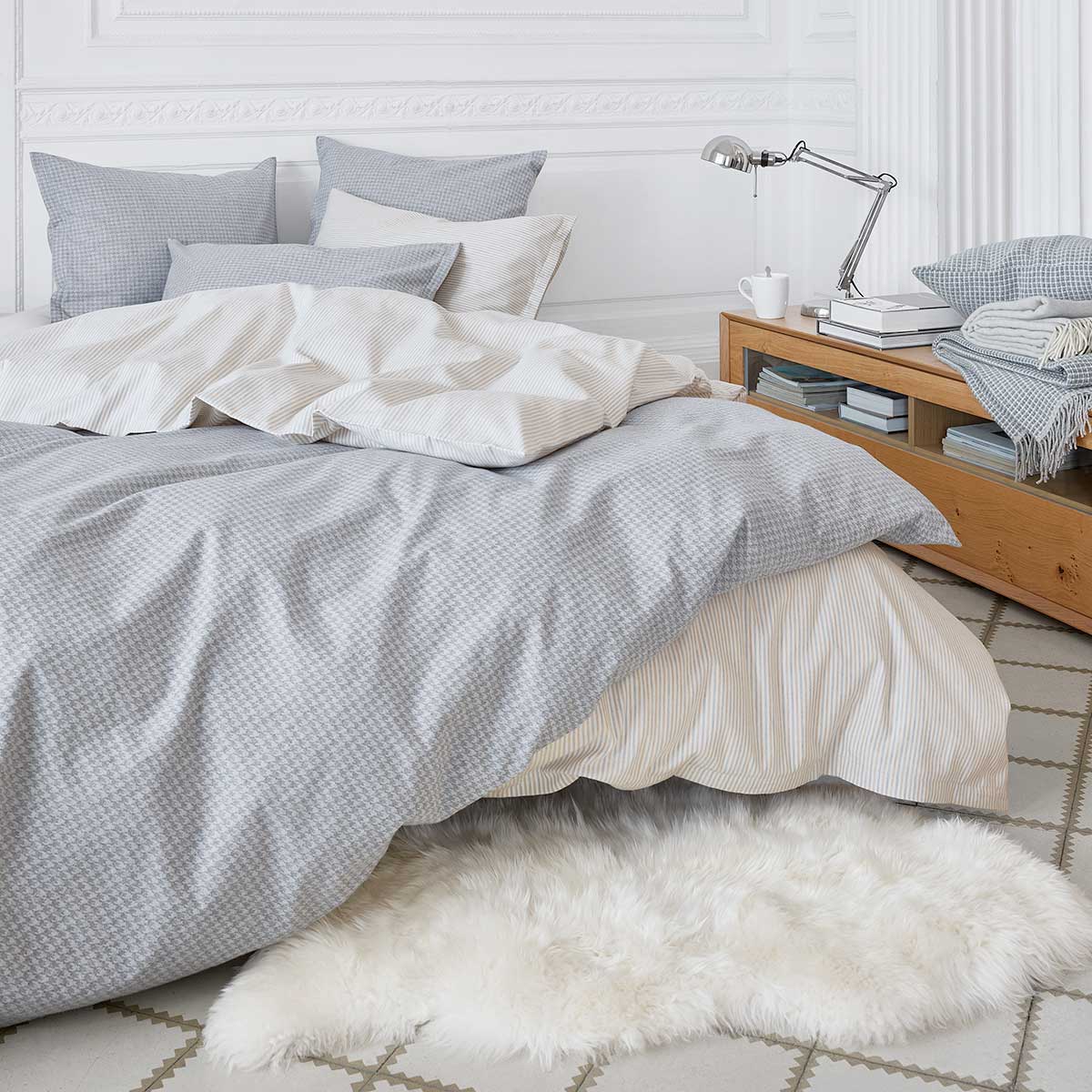 Gemütliches Schlafzimmer mit der Bettwäsche "Gustav" und grauen Akzenten Ein gemütliches Schlafzimmer dekoriert mit der Bettwäsche "Gustav" in einem feinen Streifenmuster, kombiniert mit grauen Akzenten, einem Holz-Nachttisch und einem Schaffellteppich.