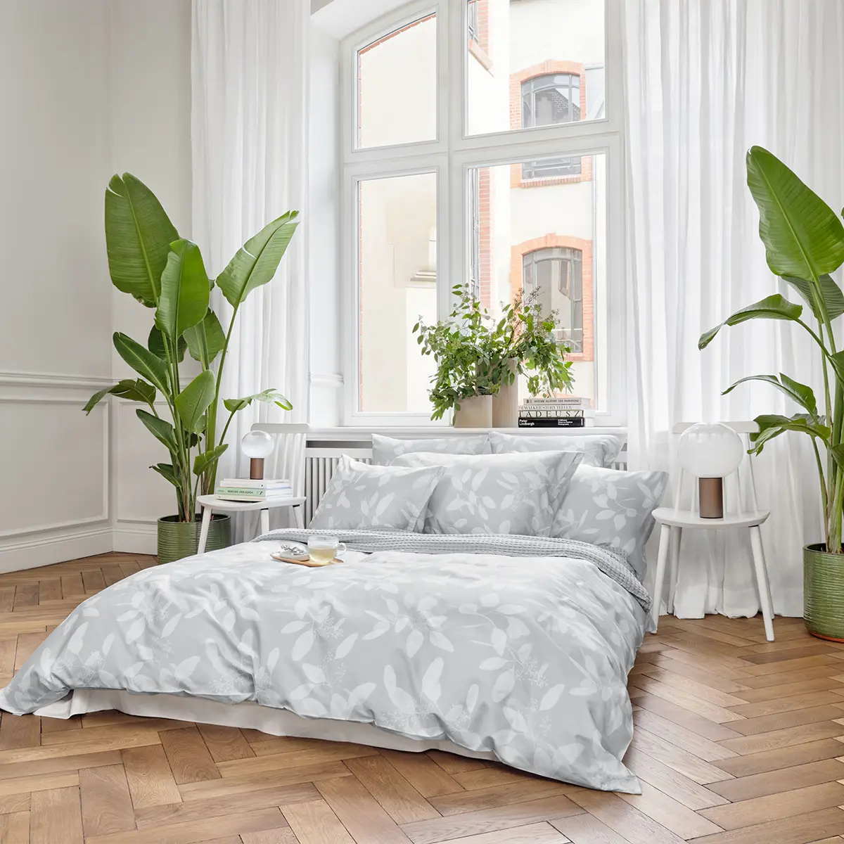 Modernes, helles Schlafzimmer mit der grauen Omeo Bettwäsche, eingerahmt von hohen Fenstern und grünen Pflanzen.