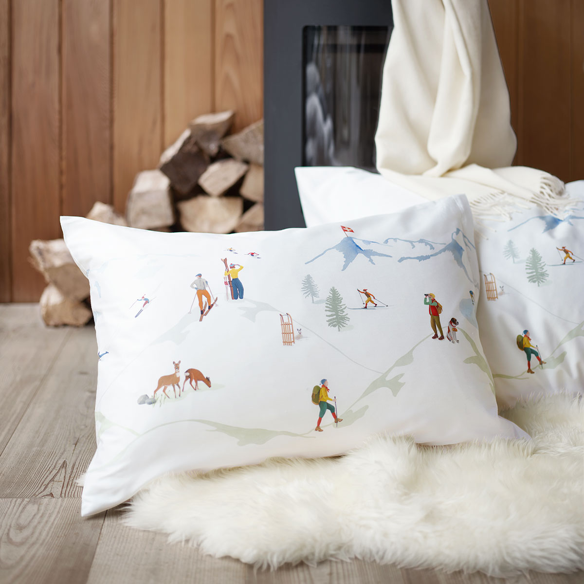 Nahaufnahme eines Kissens mit alpinem Motiv vor einem Kaminofen, umgeben von Holzscheiten und einem weissen Fellteppich.