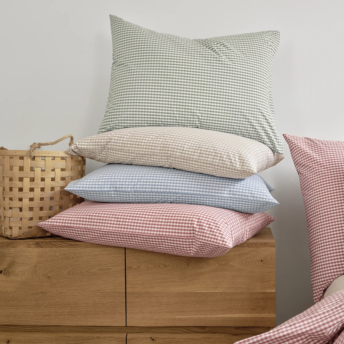 Eine Sammlung von Kissen in verschiedenen Farben der Schlossberg Bettwäsche Andrin im Karomuster: Beige, Blau, Rot und Grün. Die Kissen sind ordentlich auf einem Holzregal gestapelt und verleihen dem Raum eine frische, gemütliche Atmosphäre.