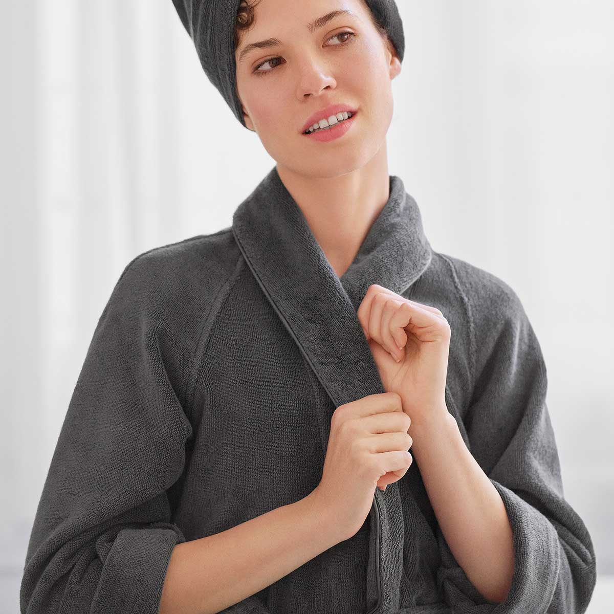 Eine Frau trägt den grauen Schlossberg Bademantel aus der Coshmere-Kollektion und einen Haarturban, während sie den Bademantel schliesst.