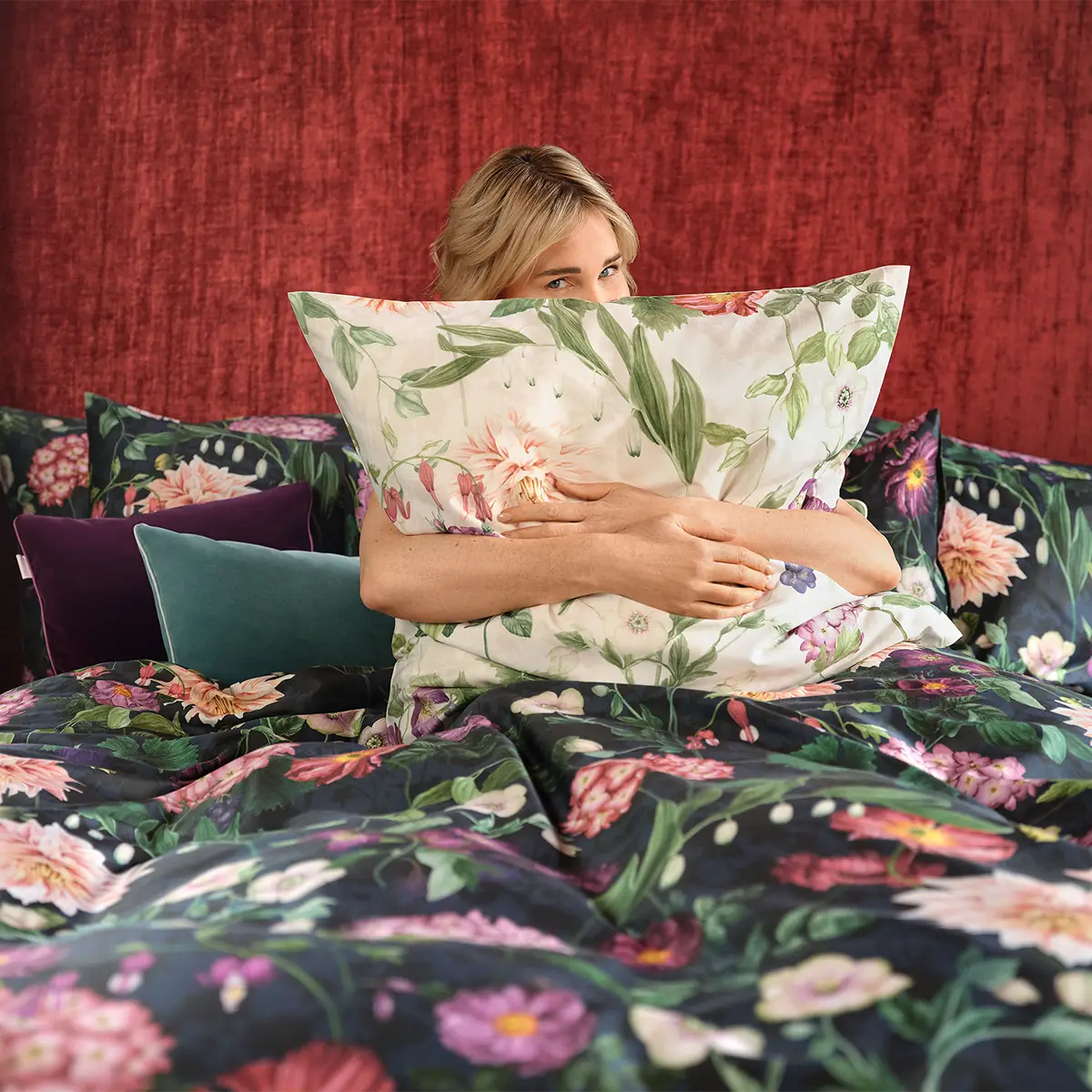 Frau mit Ophelia Kopfkissen Eine Frau umarmt ein Kopfkissen mit dem floralen Design der Ophelia Bettwäsche. Der Hintergrund ist ein tiefroter Stoff, der eine gemütliche Atmosphäre schafft.