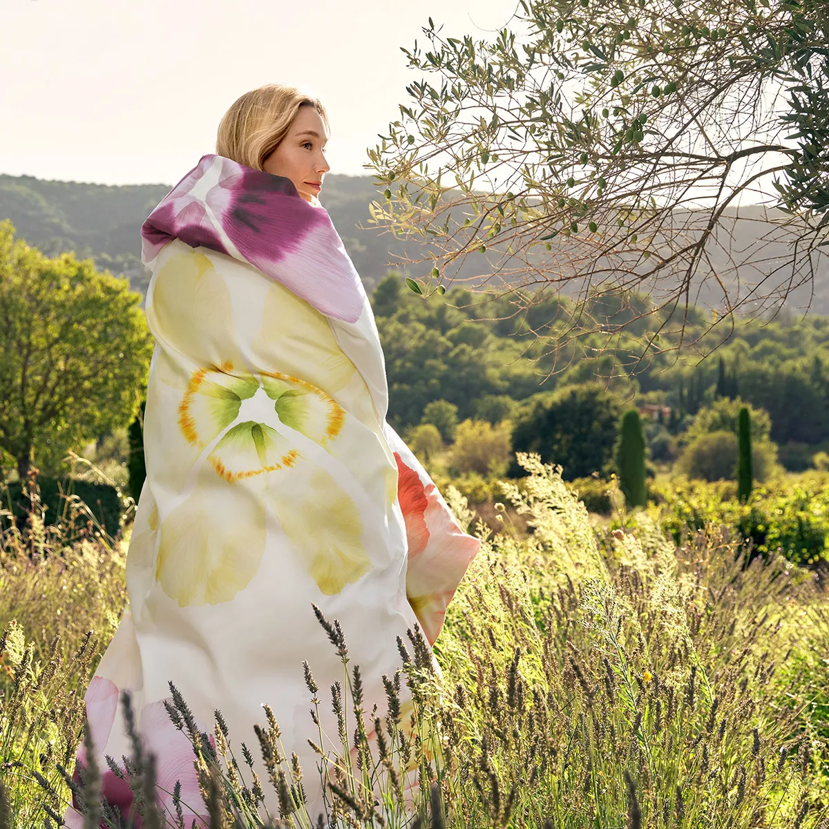 Schlossberg Bettwäsche Aurora Model im Feld von der Seite Eine blonde Frau steht inmitten einer sonnendurchfluteten Wiesenlandschaft, eingehüllt in die luxuriöse Schlossberg-Bettwäsche Aurora. Das weiche, mit floralen Blüten verzierte Design harmoniert perfekt mit der idyllischen Natur im Hintergrund.