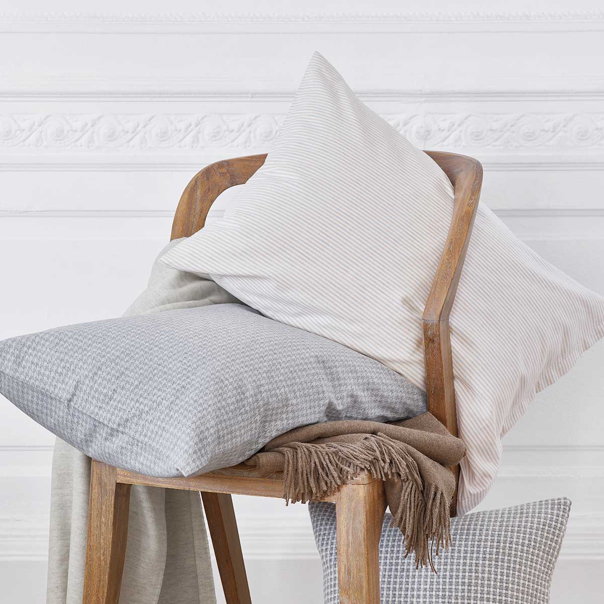 Dekorative Kissen mit der Bettwäsche "Gustav" Kissen aus der Bettwäsche "Gustav", stilvoll auf einem Holzstuhl arrangiert, mit einer Mischung aus beige-grauen Streifen und einem dezenten Muster, dekoriert mit einer weichen Decke.