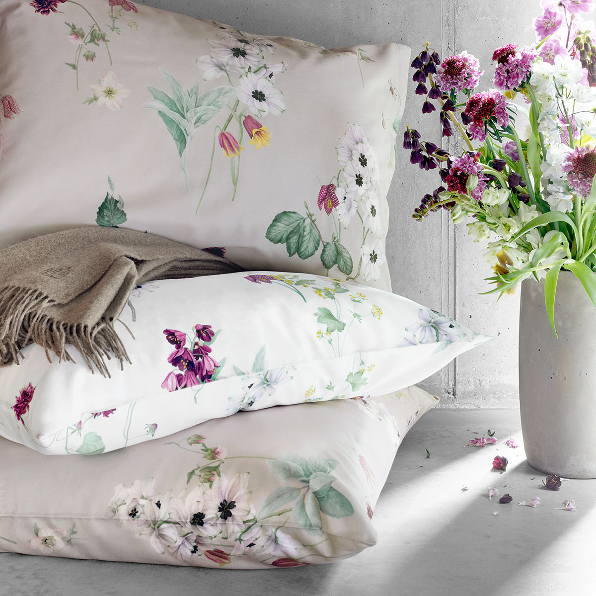 Nahaufnahme von dekorativen Kissen und Bettwäsche "Freya" mit floralen Akzenten, kombiniert mit einem Blumenstrauss in einem modernen Schlafzimmer.
