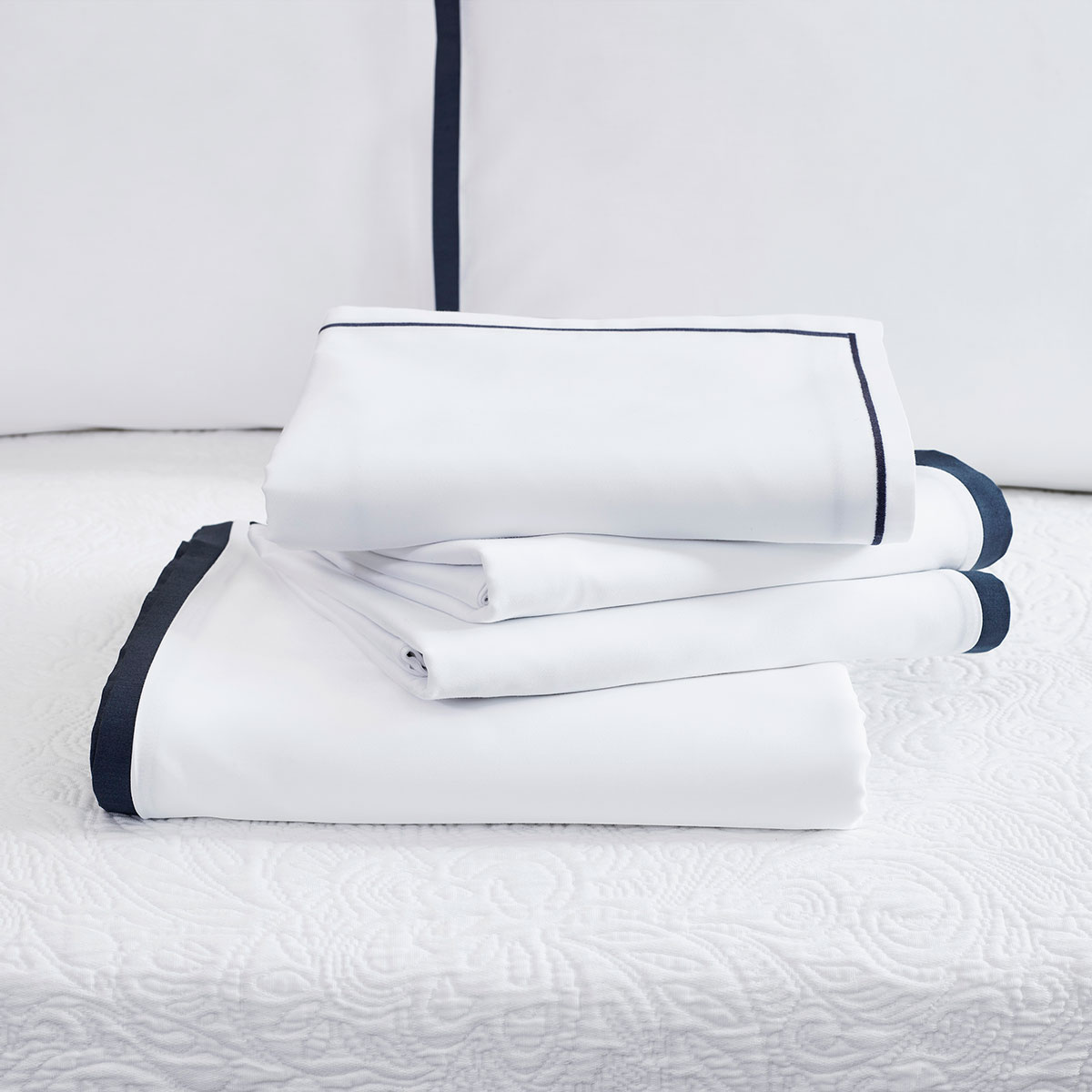 Ein Stapel der Bettwäsche Clark in weiss mit dunklem Rand, auf einem Bett drapiert.