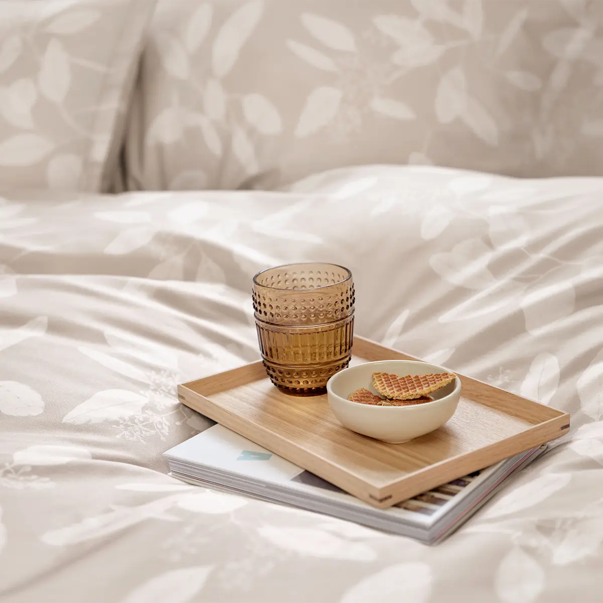 Nahaufnahme der Omeo Bettwäsche in beige mit dezentem Blätterdesign, darauf ein Tablett mit einem Glas und einem Schälchen Gebäck.