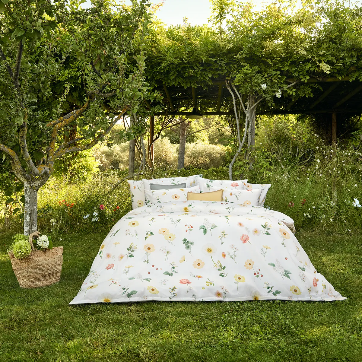 Ein Bett mit Schlossberg Bettwäsche Zinnia steht mitten in einem idyllischen Garten unter einer grünen Pergola. Die Bettwäsche zeigt ein florales Design mit gelben, rosafarbenen und weißen Blüten sowie zarten grünen Blättern auf einem weißen Hintergrund.