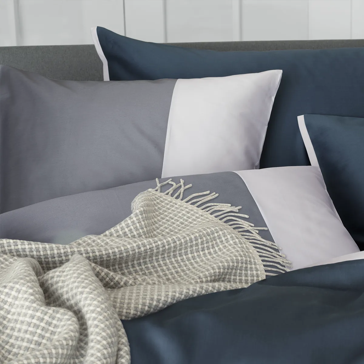 Schlossberg Bettwäsche Allen Acier Glace Ein modernes Bett mit Schlossberg Bettwäsche im Allen Design, das sich aus dunkelblauen und grauen Kissenbezügen zusammensetzt, begleitet von einem weichen, karierten Überwurf.