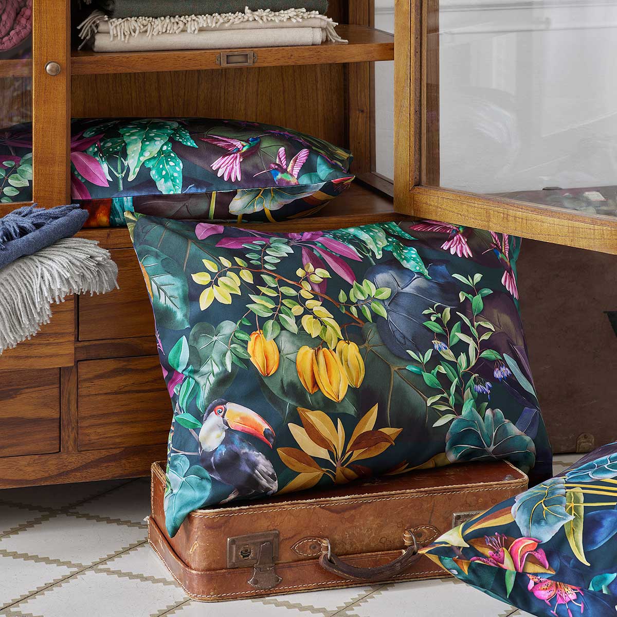 Kissen aus der Jane-Bettwäsche Kollektion mit exotischem Tropenmuster in kräftigen Farben, präsentiert auf einem Holzschrank.