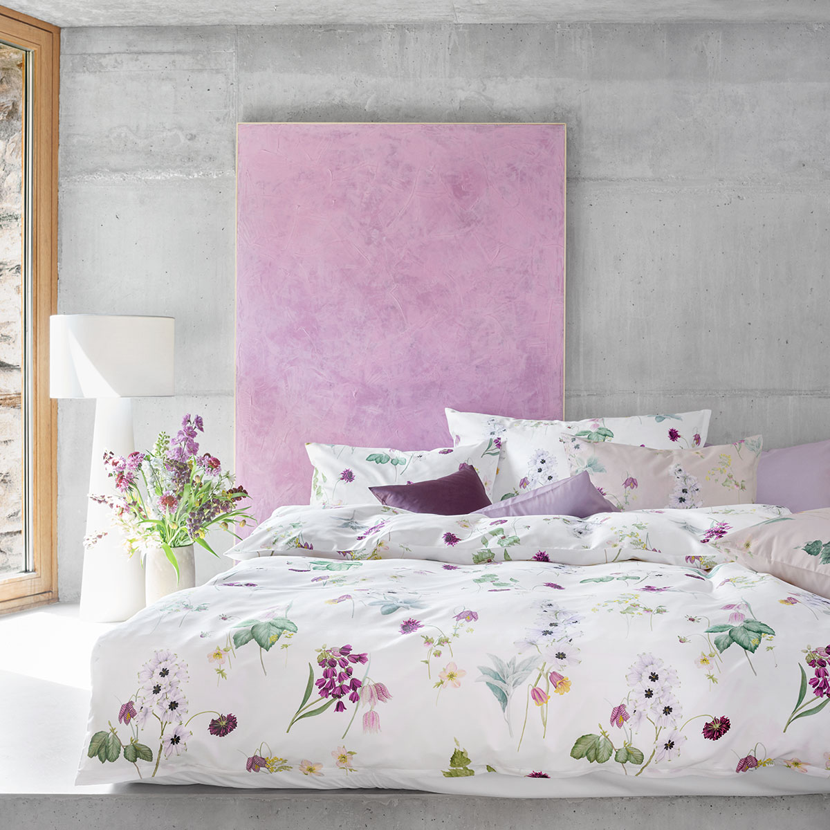 Ein modernes Schlafzimmer mit weisser Bettwäsche "Freya", dekoriert mit floralen Mustern und pastellfarbenen Kissen, in einem Raum mit Betonwänden und einer rosa Wanddekoration.