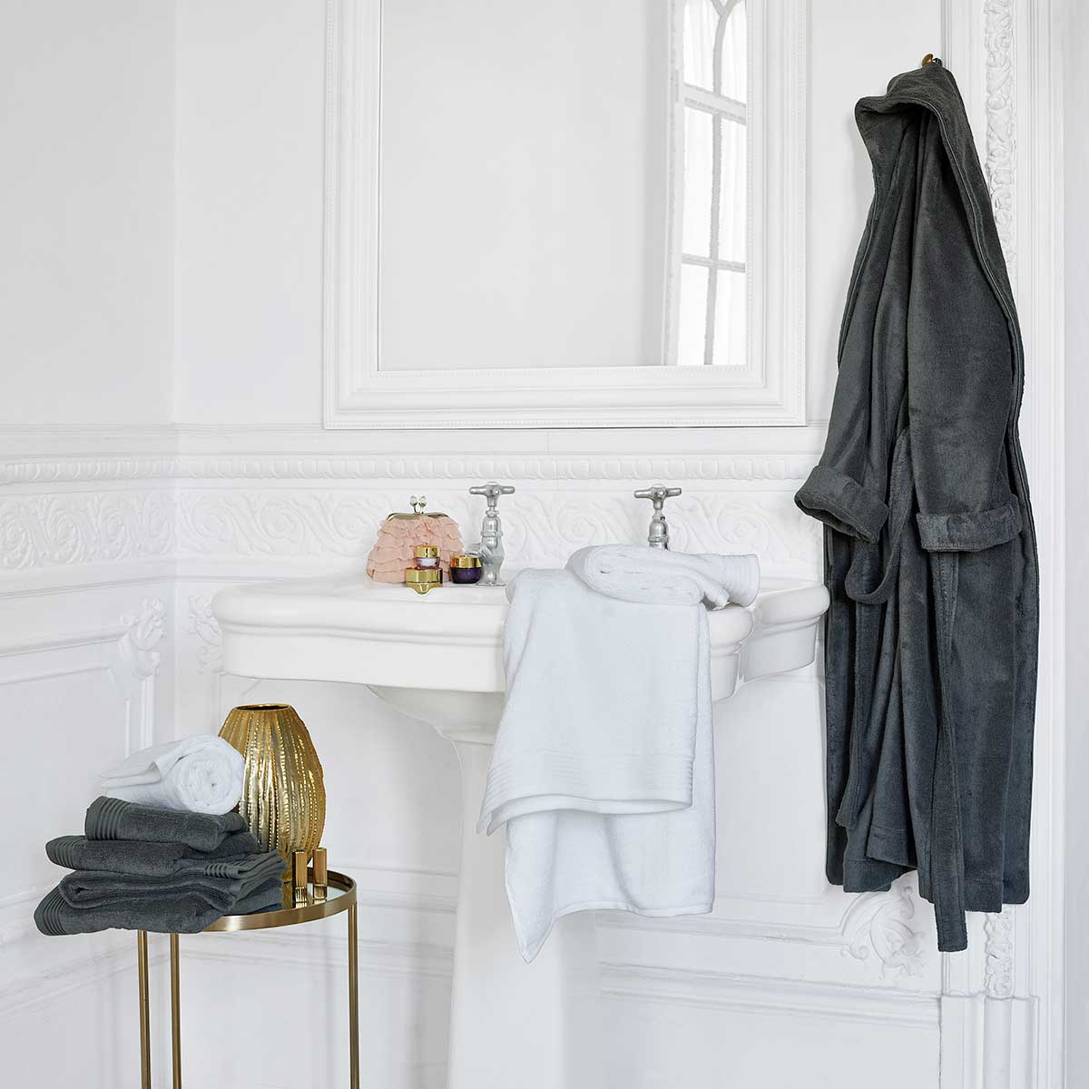 Ein Badezimmer mit einem grauen Schlossberg Frottier Coshmere Bademantel an einem Haken, weissen Handtüchern auf einem Tisch und weiteren Pflegeprodukten.